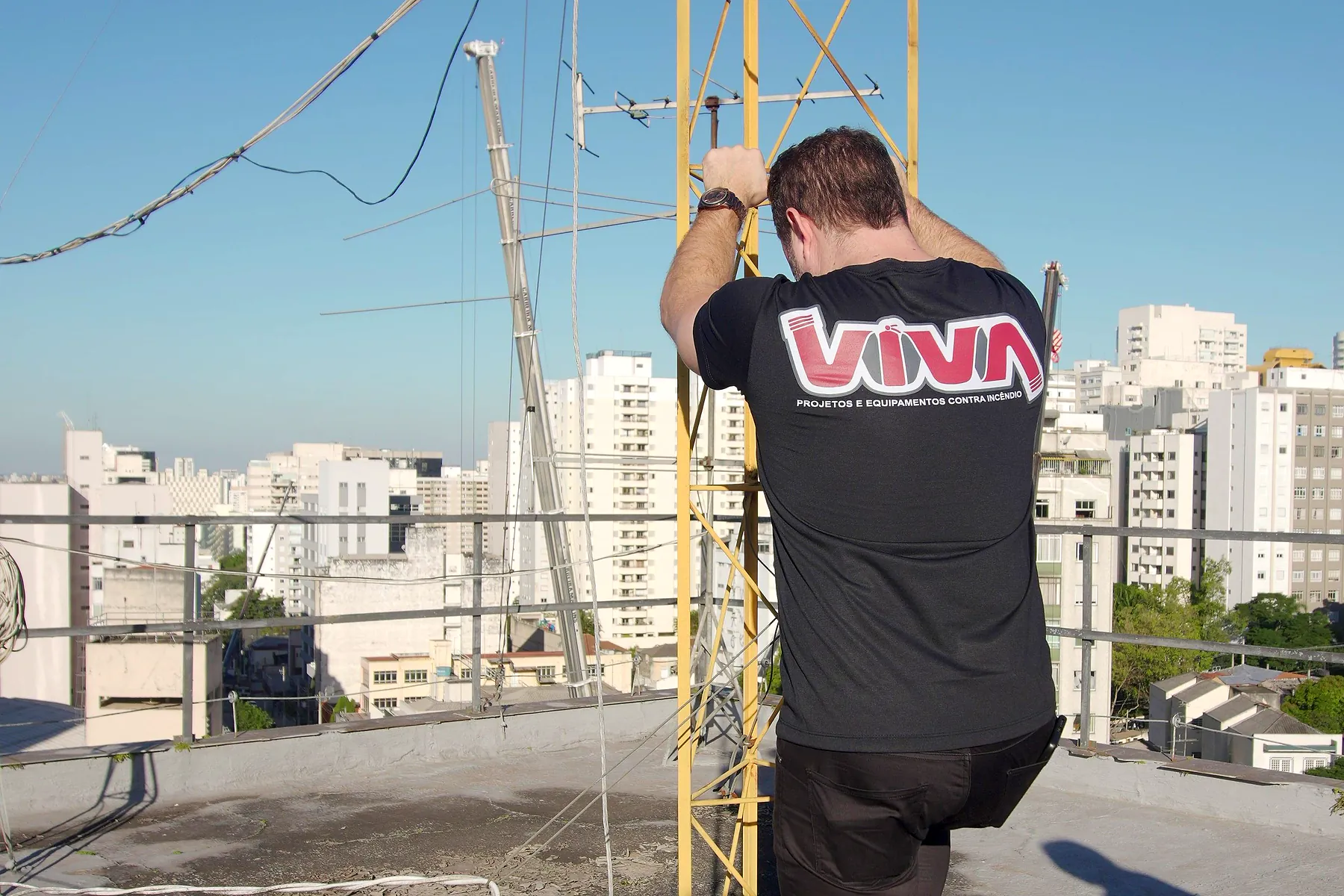 Engenheiro da VIVA de costas, trabalhando na torre de SPDA em cobertura, com a cidade ao fundo